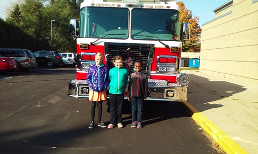 Three girls stand in front of a fire truck on a sunny day.